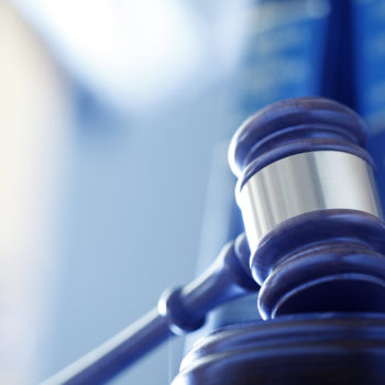 A wooden gavel rests on its sounding block in front of a row of law books and an office interior that are out of focus in the background. Photographed using a shallow depth of field.